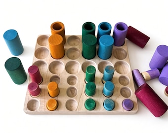 May include: A square wooden board with a grid of circular indentations, featuring various cylindrical wooden pieces in shades of blue, green, purple, orange, and red placed inside the holes or scattered around. The text reads Grapat is sold separately.