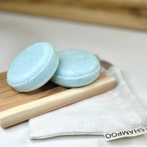 May include: Two light blue, round shampoo bars rest on a wooden soap dish, next to a small white bag labeled "SHAMPOO". The bars have a slightly textured surface and a rounded edge. The background is a light brown.