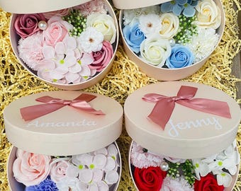 May include: Four round gift boxes with lids, each containing a different arrangement of artificial flowers. The boxes are pink and have a satin ribbon tied around them. The flowers are made of soap and are in various colors, including pink, white, blue, and red. The boxes are labeled with the names "Amanda" and "Jenny".