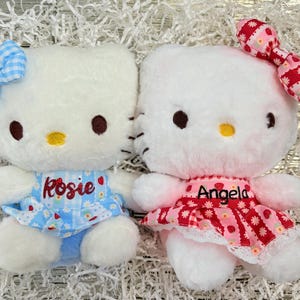 May include: Two plush white cat dolls with embroidered facial features. One doll wears a blue dress with the name "Rosie" and a bow with a strawberry. The other doll wears a red dress with the name "Angela" and a red bow.