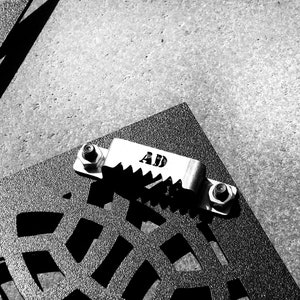 May include: A black metal grate with a silver metal clamp attached to it. The clamp has the letters "AD" engraved on it.