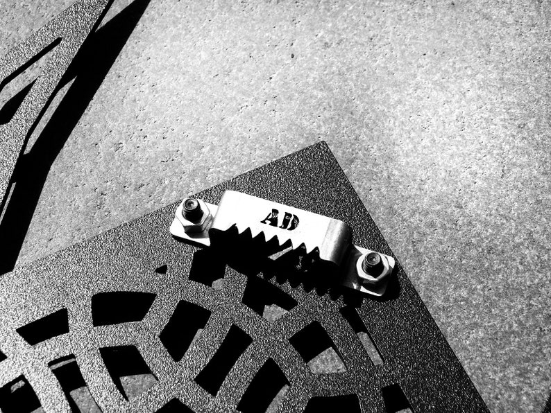 May include: A black metal grate with a silver metal clamp attached to it. The clamp has the letters "AD" engraved on it.