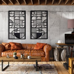 May include: Two black metal wall sculptures with geometric designs, hanging on a concrete wall above a brown leather sofa with orange throw pillows. A wooden coffee table sits in front of the sofa, and a lamp with a brown lampshade sits on a wooden desk in the background.