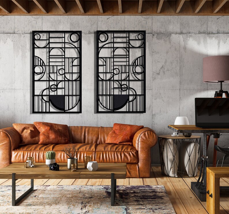 May include: Two black metal wall sculptures with geometric designs, hanging on a concrete wall above a brown leather sofa with orange throw pillows. A wooden coffee table sits in front of the sofa, and a lamp with a brown lampshade sits on a wooden desk in the background.