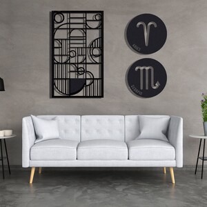 May include: A white sofa with three cushions sits in front of a grey wall with black metal wall art. The wall art features geometric shapes and two round metal signs with zodiac symbols. The signs read "Aries" and "Scorpio".
