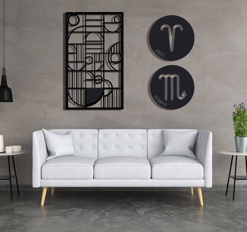 May include: A white sofa with three cushions sits in front of a grey wall with black metal wall art. The wall art features geometric shapes and two round metal signs with zodiac symbols. The signs read "Aries" and "Scorpio".