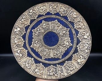May include: A round, silver-colored decorative plate with a deep blue center. The plate features intricate relief carvings of animals and floral patterns around the edge and center. The plate is likely made of metal and has an antique appearance.