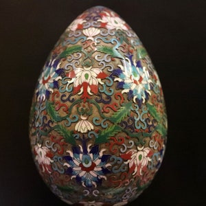 May include: An egg-shaped decorative object with a detailed floral pattern. The design features white, red, blue, and green flowers and leaves on a bronze-colored background. The egg is likely made of metal and enamel.
