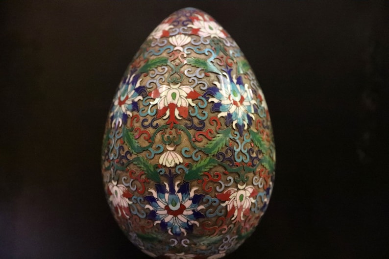 May include: An egg-shaped decorative object with a detailed floral pattern. The design features white, red, blue, and green flowers and leaves on a bronze-colored background. The egg is likely made of metal and enamel.
