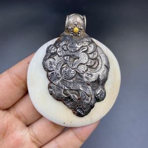May include: A round, off-white pendant with a detailed silver metal lion design. The pendant has a silver loop at the top with a small yellow stone. The pendant is held in a hand.