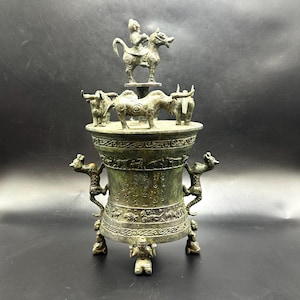 Puede incluir: Jarrón decorativo de color bronce con detalles intrincados. Presenta un jinete a caballo, varias figuras de animales y patrones ornamentados. El jarrón está sostenido por tres figuras y tiene asas con forma de dragón.