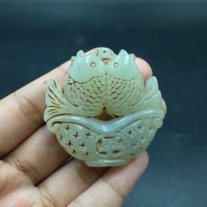 Puede incluir: Colgante de jade verde pálido tallado con dos peces enfrentados. Los peces están detallados con escamas y aletas, y están colocados dentro de un cuenco decorativo con formas de estrella recortadas y un símbolo central.