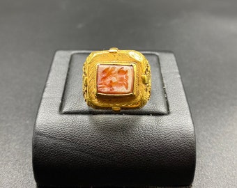 May include: A gold ring with a square, reddish-orange stone featuring a carved design. The ring has an ornate, detailed setting with decorative elements. The ring is displayed on a black surface.