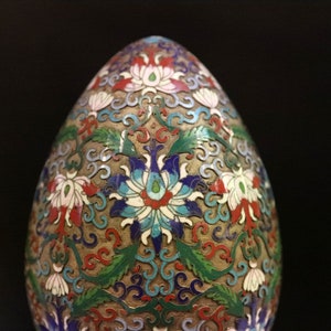 May include: An egg-shaped decorative object with a detailed floral pattern. The design features white, red, blue, and green flowers and leaves on a gold-colored background. The object has a smooth, rounded shape.