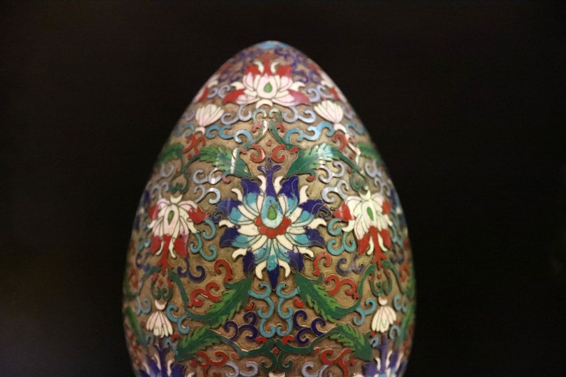 May include: An egg-shaped decorative object with a detailed floral pattern. The design features white, red, blue, and green flowers and leaves on a gold-colored background. The object has a smooth, rounded shape.
