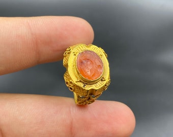 May include: A gold ring with an oval, orange-red stone featuring a carved face. The ring has intricate gold filigree and decorative elements around the stone and band. The ring is held between fingers.