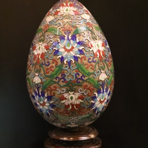May include: An ornate, egg-shaped decorative object with a floral design in red, white, blue, and green. The egg sits on a small, dark wooden stand. The intricate pattern covers the entire surface.