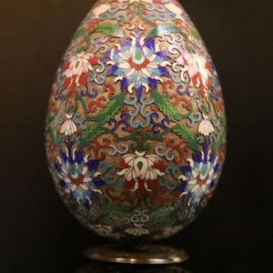 May include: An egg-shaped decorative object with a detailed floral design. The object features a base and is adorned with red, white, blue, and green floral patterns on a gold background. The intricate design suggests a handcrafted item.