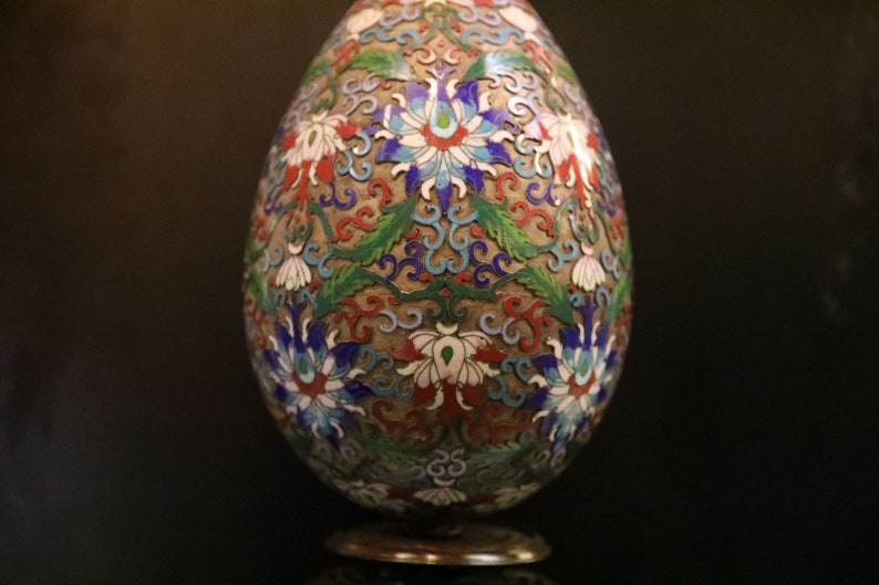 May include: An egg-shaped decorative object with a detailed floral design. The object features a base and is adorned with red, white, blue, and green floral patterns on a gold background. The intricate design suggests a handcrafted item.