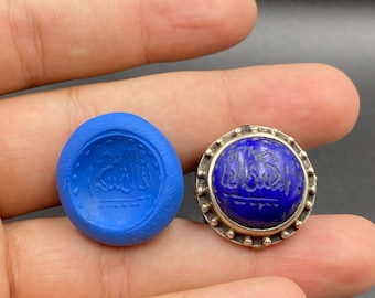 Può includere: Un anello argentato con una grande pietra blu scuro con un disegno inciso. Accanto, un'impronta blu dello stesso disegno. L'anello e l'impronta sono tenuti in una mano. Lo sfondo è scuro.