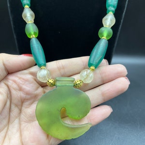 Puede incluir: Un collar de vidrio verde con un colgante en forma de ola. El collar presenta cuentas verdes, transparentes y doradas. El colgante es de color verde claro y tiene un aspecto esmerilado.