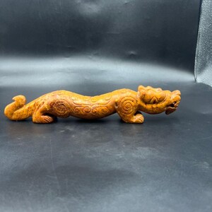 Puede incluir: Una escultura de madera tallada de un dragón estilizado en posición horizontal. El dragón es de color marrón claro cálido con detalles intrincados, incluyendo patrones en espiral a lo largo de su cuerpo y una cabeza detallada. La escultura está sobre un fondo oscuro.