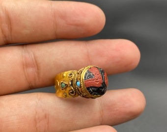 May include: A gold ring with a rectangular, red and black stone. The ring has turquoise and gold accents. The ring is held in a hand against a dark background.