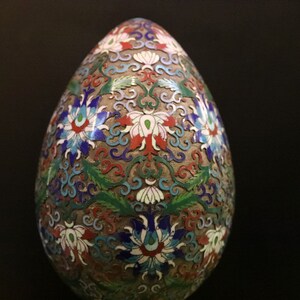 May include: An egg-shaped decorative object with a detailed floral design in red, white, blue, and green. The egg has a metallic, possibly cloisonn&eacute;, appearance with intricate patterns and a dark background.