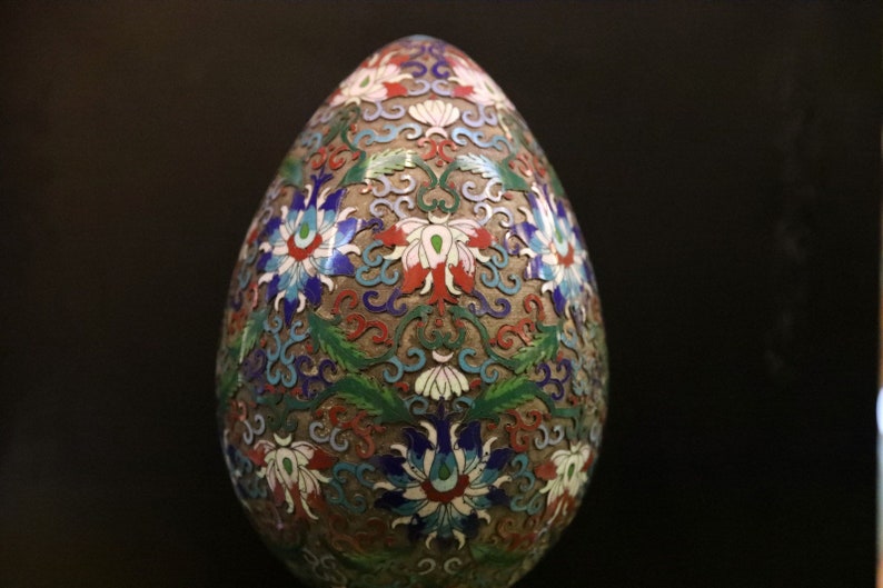 May include: An egg-shaped decorative object with a detailed floral design in red, white, blue, and green. The egg has a metallic, possibly cloisonn&eacute;, appearance with intricate patterns and a dark background.
