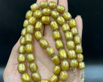 May include: A strand of golden, barrel-shaped beads. The beads are strung together with a natural-colored cord. The beads have a metallic, reflective surface. The background is a dark, solid color.