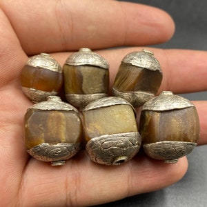 Könnte beinhalten: Sechs ovale Perlen mit silberfarbenen Metallkappen. Die Perlen haben ein durchscheinendes, braun-beiges Streifenmuster. Die Metallkappen weisen ein geätztes Design auf. Diese Perlen können für die Schmuckherstellung verwendet werden.