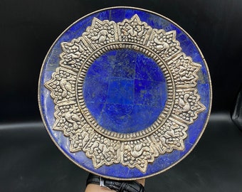May include: A round, decorative plate with a deep blue, mosaic-style center. The plate features a silver-colored, ornate border with repeating floral and bird designs. The plate is held against a dark background.