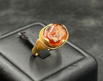 May include: A gold ring with an oval, reddish-orange stone. The stone features a detailed carving of a face or figure. The ring has a textured band and a simple, elegant design. The ring is displayed on a black surface.
