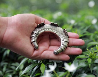 May include: A silver-colored cuff bracelet with intricate embossed designs and floral accents. The open-ended bracelet is held in a hand against a backdrop of green foliage, showcasing its ornate details and craftsmanship.