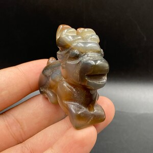Pode incluir: Estatueta de pedra esculpida de uma criatura mítica, possivelmente um Qilin. A escultura apresenta tons castanhos e bege, com detalhes precisos e um acabamento polido. A criatura é retratada em posição sentada.