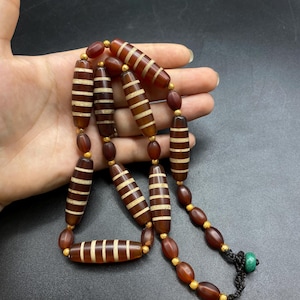 Può includere: Una collana con perline marroni allungate a strisce bianche orizzontali, alternate a piccole perline marroni rotonde e minuscoli distanziatori dorati. Il filo è nero e presenta una piccola perla verde in corrispondenza della chiusura.