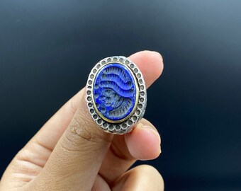 Può includere: Anello ovale con una pietra blu scuro con motivi scolpiti a forma di onde. La pietra è incastonata in una fascia argentata con un bordo interno dorato e un bordo a puntini. Un gioiello.