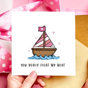 May include: A square greeting card featuring a cartoon sailboat. The boat has a pink and white striped sail and a pink heart flag. The text "YOU REALLY FLOAT MY BOAT" is below the boat. Small pink hearts are scattered around the boat.