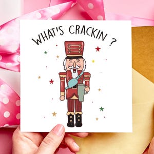 May include: A greeting card with a cartoon nutcracker soldier holding a mug. The card reads "WHAT'S CRACKIN ?" in black. The nutcracker is in a red uniform with a blue strap and is surrounded by colourful stars.