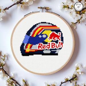 May include: A cross-stitch of a Red Bull racing helmet, with the brand's logo and colours. The design is set within a wooden embroidery hoop, surrounded by white fabric and floral accents. The helmet design includes blue, red, yellow, and white.