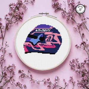 May include: A cross-stitch design in a wooden hoop. The design features a stylized, pixelated image in shades of blue, pink, and purple. The background is white, and the hoop is surrounded by pink flowers on a pink background.