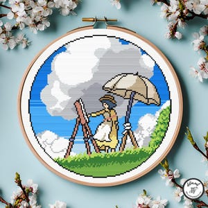 Puede incluir: Un bastidor de bordado de punto de cruz con una imagen pixelada de una persona pintando al aire libre. La escena incluye un cielo azul, nubes, hierba verde y un paraguas beige. El bordado está en un bastidor de madera.