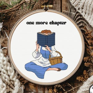 May include: A circular wooden embroidery hoop containing a cross-stitch design of a person sitting cross-legged while reading a blue book. The text "one more chapter" is stitched in black above the figure, who wears a blue dress and apron.