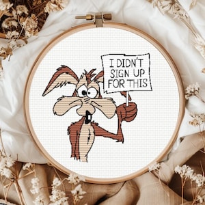 May include: A cross-stitch embroidery in a round wooden hoop featuring a brown coyote character with wide eyes holding a white sign that reads I DIDN'T SIGN UP FOR THIS in black capital letters against a white fabric background.
