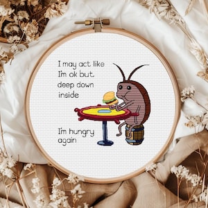 Puede incluir: Un bordado de punto de cruz en un bastidor de madera con una cucaracha marrón sentada en un barril en una mesa, sosteniendo una hamburguesa. El texto dice: I may act like I'm ok but deep down inside I'm hungry again. Diseño sobre tela blanca.
