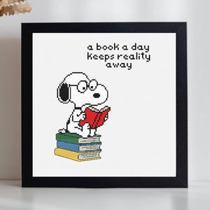 May include: A framed cross-stitch embroidery featuring a white dog with black ears wearing glasses, sitting on a stack of three books colored green, blue, and yellow, while reading a red book. The text reads: a book a day keeps reality away.