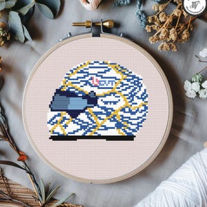 May include: A cross-stitch embroidery hoop featuring a pixelated design of a racing helmet. The helmet is primarily blue and white with yellow accents, and the word "OUT" is visible. The hoop is set against a light pink fabric, with floral accents.
