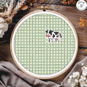 Può includere: Un telaio da ricamo circolare in legno contenente un motivo a punto croce di una mucca bianca e nera centrata su una griglia geometrica floreale ripetuta verde e gialla, con un piccolo muso e una mammella rosa.