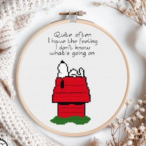 May include: A cross-stitch embroidery hoop featuring Snoopy on his red doghouse with the quote "Quite often I have the feeling I don't know what's going on." The design is set against a white background, with a wooden hoop and a cozy, off-white knitted blanket.