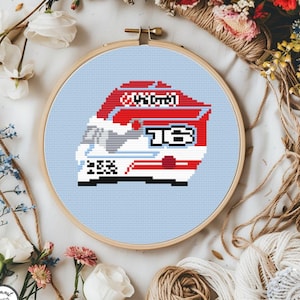 May include: A cross-stitch embroidery hoop featuring a pixelated design of a race car. The car is primarily red and white, with the number "13" prominently displayed. The background is light blue, and the hoop is surrounded by flowers and twine.